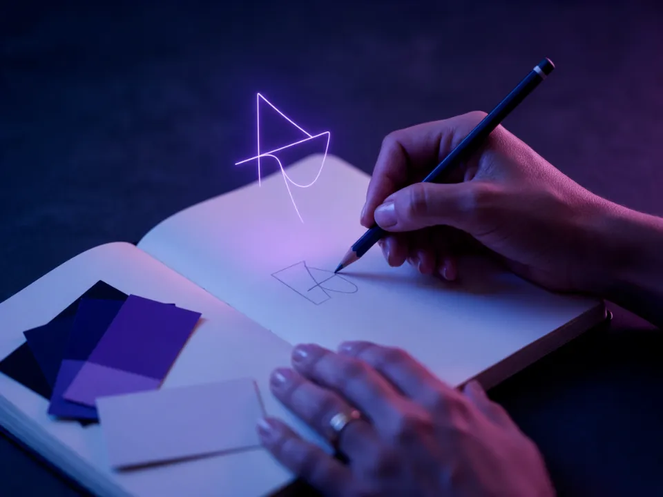 Mãos desenham o primeiro símbolo de uma marca num caderno, enquanto o mesmo símbolo flutua em luz roxa, ao lado de amostras de cor